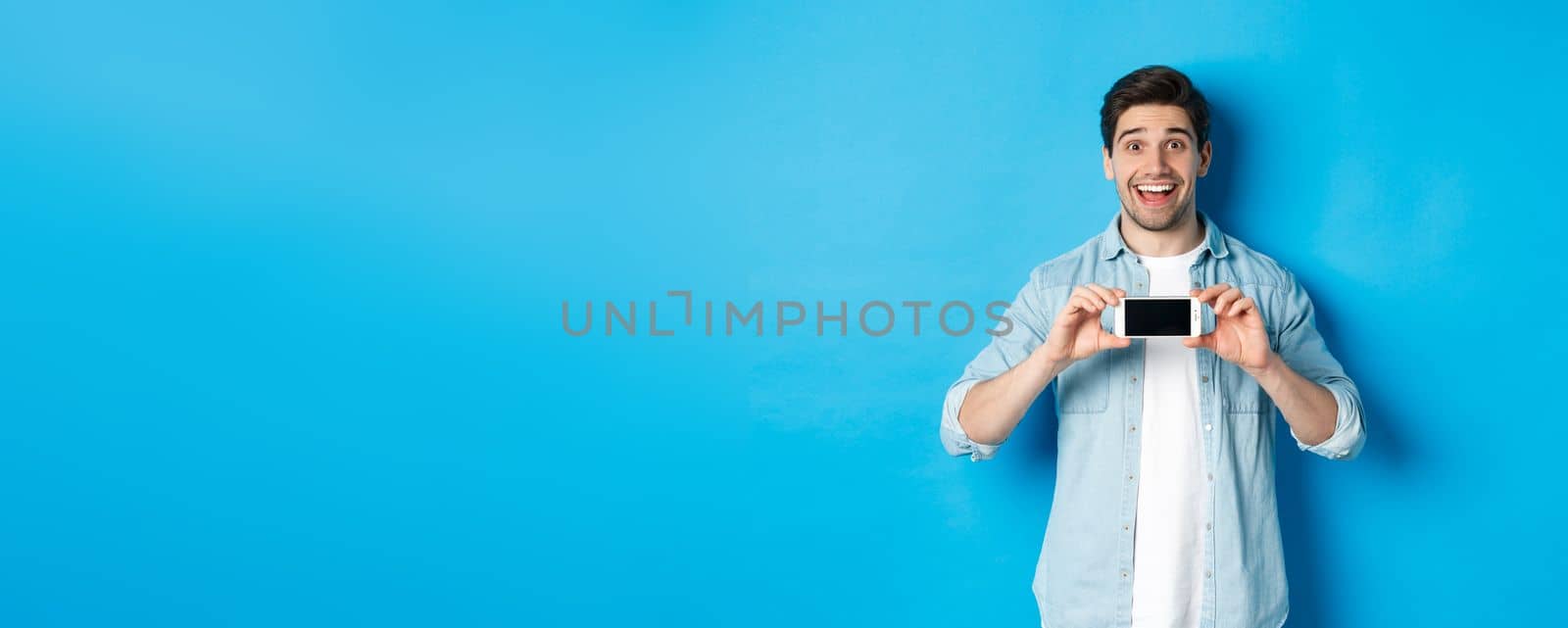 Amazed smiling man showing smartphone screen, internet promo offer, standing against blue background.