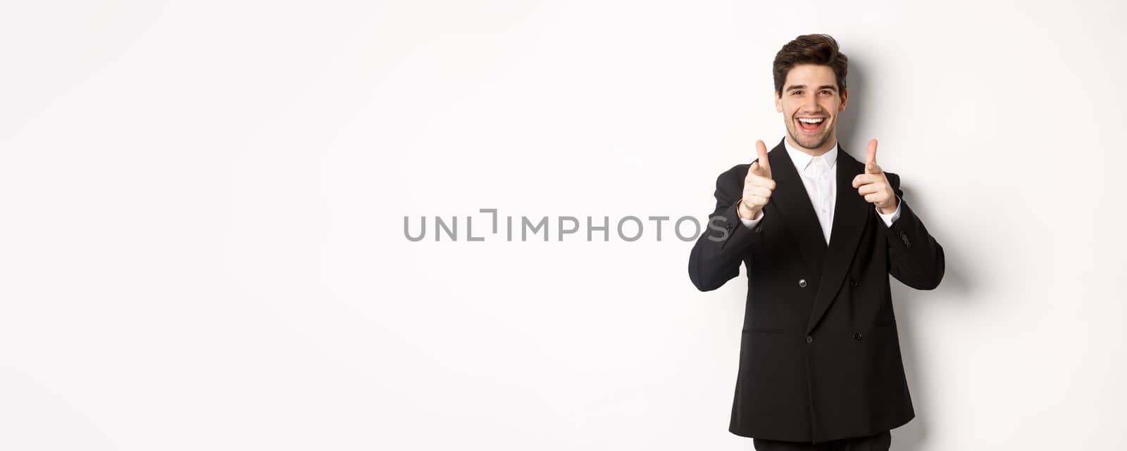 Concept of new year party, celebration and lifestyle. Handsome and successful guy in black suit, pointing fingers at camera and congratulating you, standing over white background.