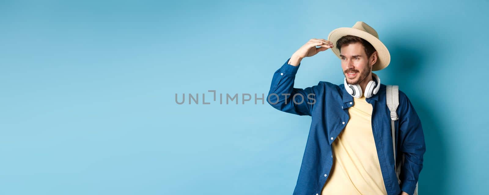 Handsome tourist in straw hat, covering face from sun and looking at distance aside, smiling pleased, travelling on summer vacation, standing over blue background by Benzoix