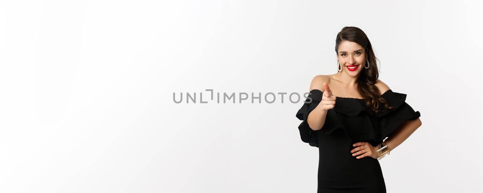 Fashion and beauty. Sassy pretty woman in black dress and makeup, pointing finger at camera to congratulate or praise, standing over white background by Benzoix