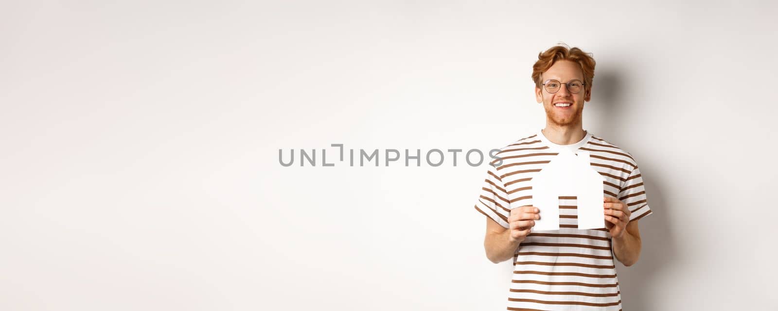 Handsome young man with beard and red hair, wearing glasses and striped t-shirt, showing paper house cutout and smiling, concept of real estate and buying property by Benzoix