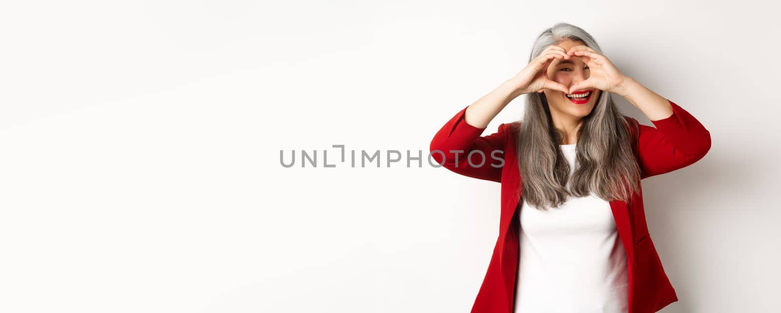 Lovely asian middle-aged woman with grey hair, wearing red blazer, showing heart sign and peeking throught it, I love you gesture, standing over white background.