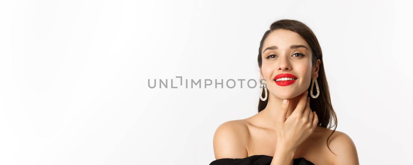 Fashion and beauty concept. Close-up of gorgeous brunette woman with red lips, touching face and smiling self-assured, standing over white background.