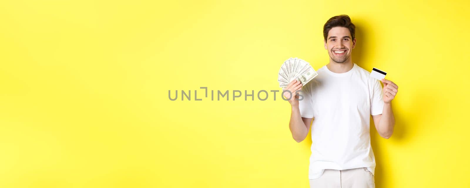 Young man withdraw money from credit card, smiling pleased, standing over yellow background.