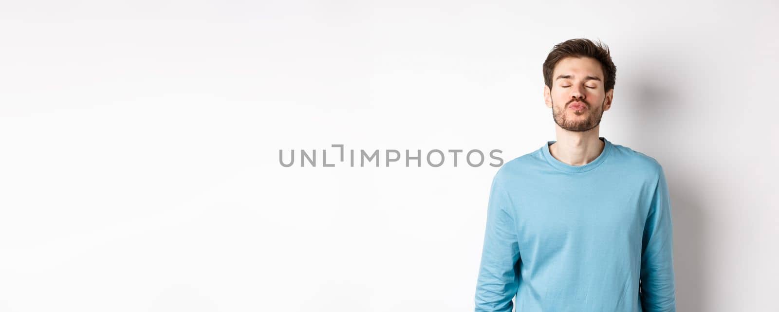 Attractive boyfriend waiting for kiss with puckered lips and closed eyes, standing in sweatshirt over white background by Benzoix