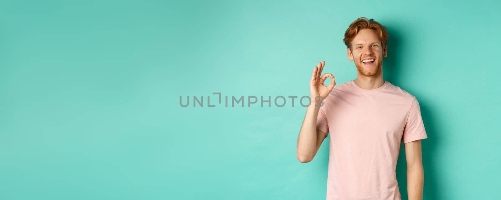 Handsome young bearded man in t-shirt showing Ok sign, smiling with white teeth and saying yes, agree with you, standing over turquoise background.