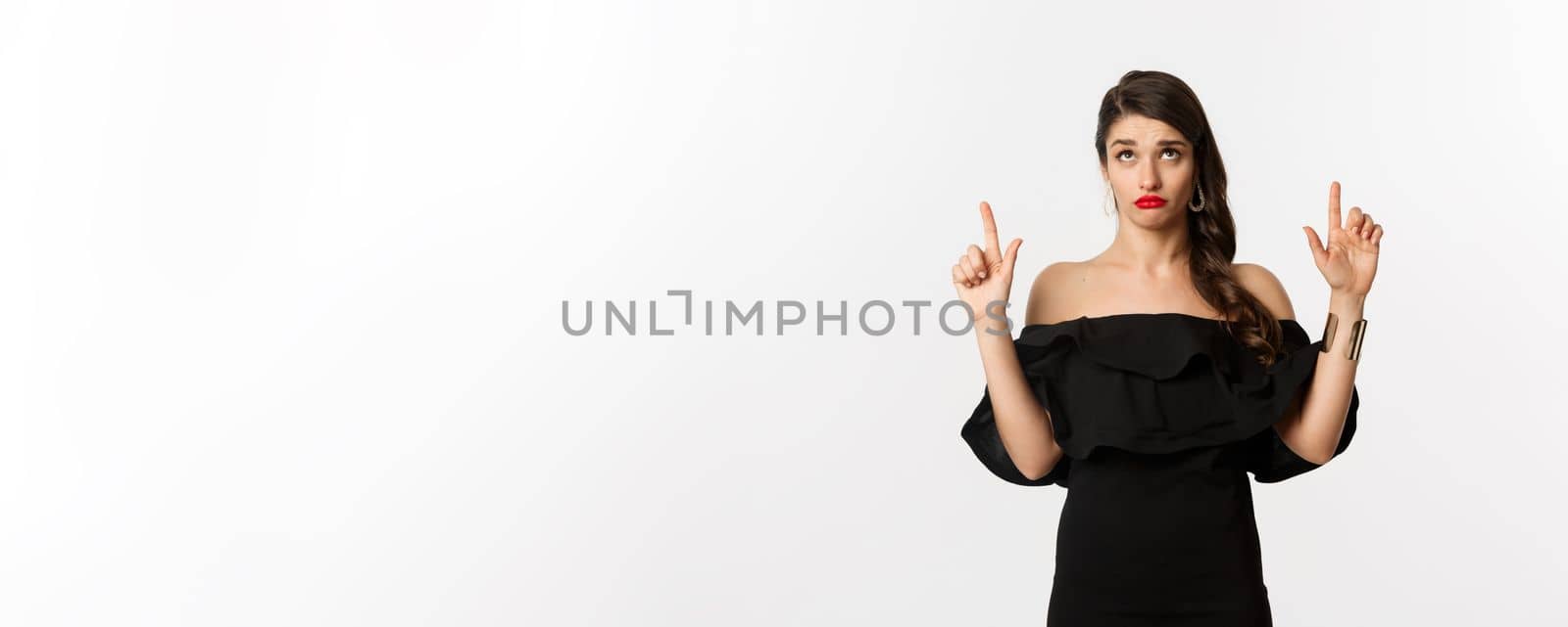 Fashion and beauty. Silly woman in black dress, red lips, looking and pointing fingers up with unamused doubtful expression, white background.