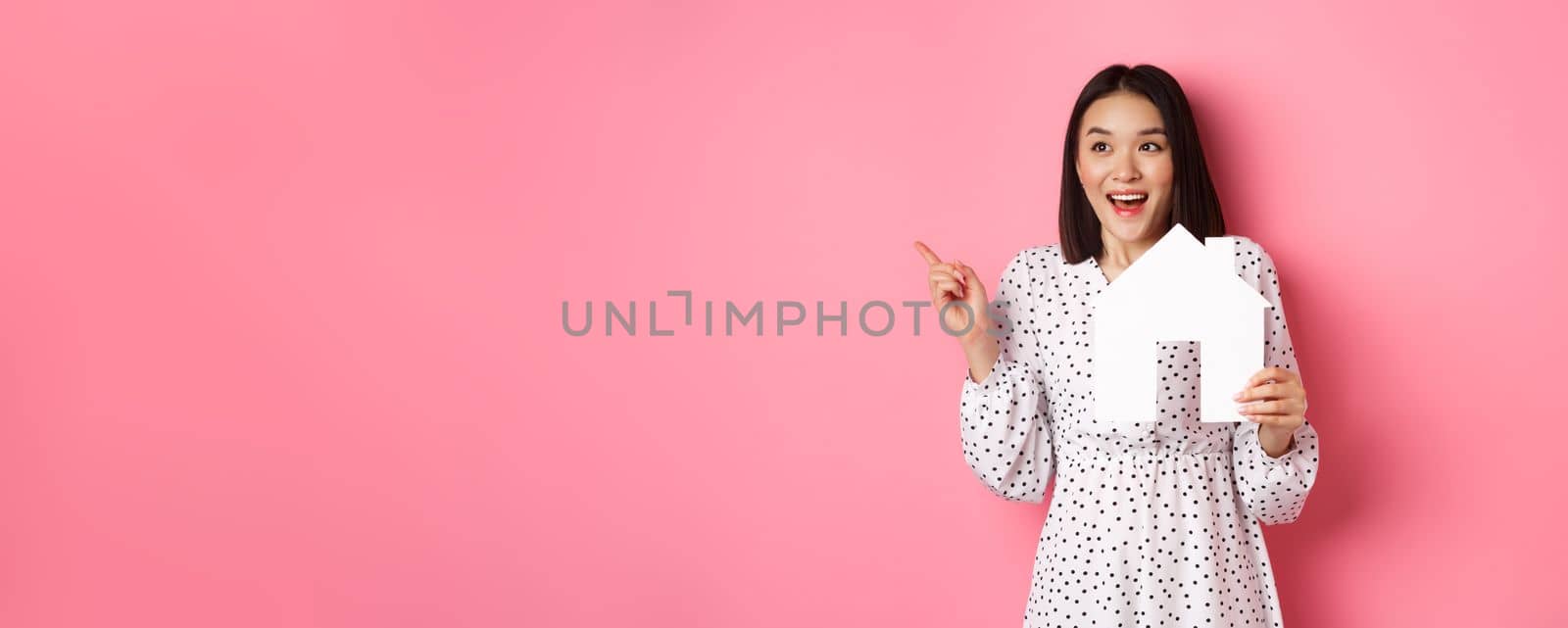 Real estate. Excited asian woman showing paper house model, pointing and looking left at copy space, standing over pink background by Benzoix