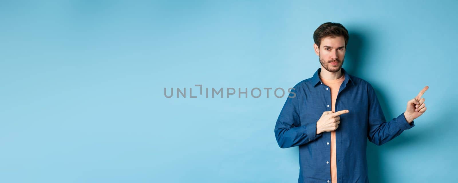 Handsome bearded man pointing fingers rights at empty space and looking confident, standing on blue background.