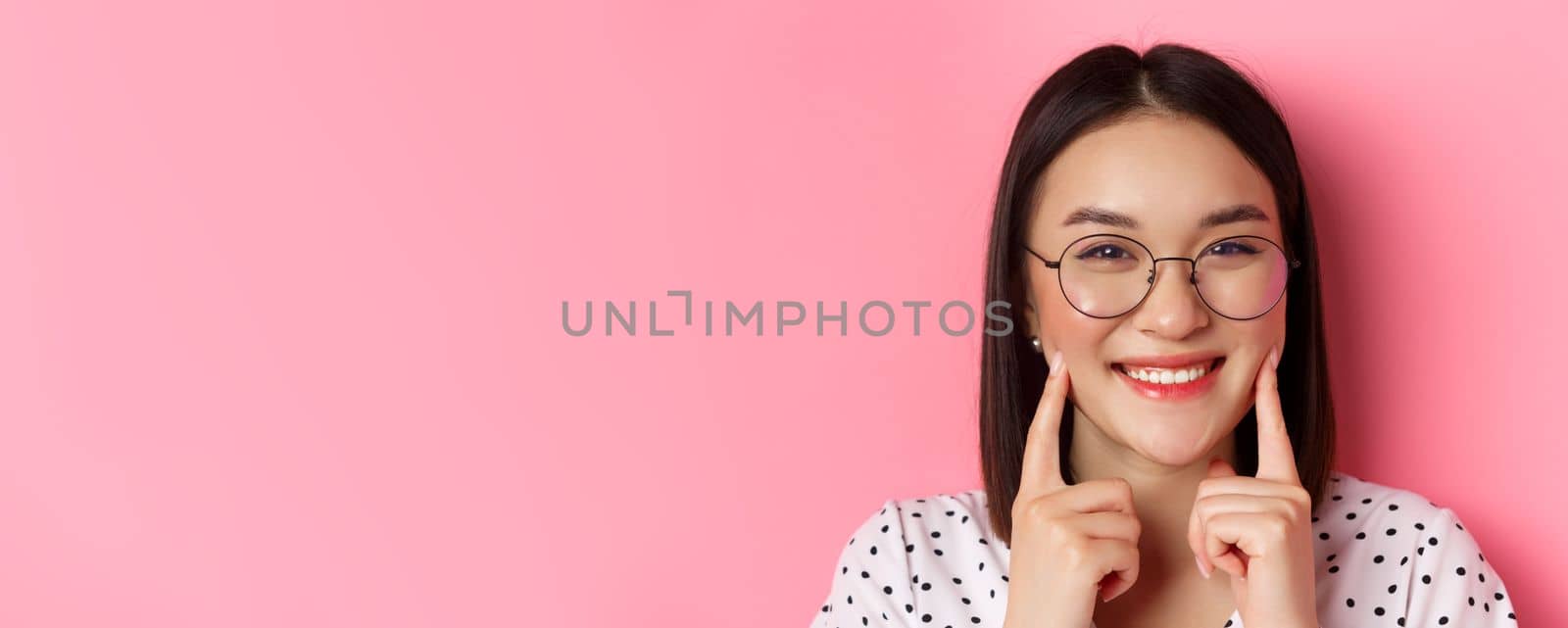 Beauty concept. Headshot of adorable asian girl in trendy glasses smiling, poking cheeks and showing cute dimples, standing over pink background.