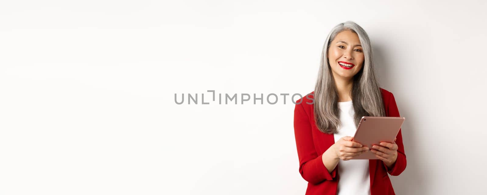 Business. Successful senior businesswoman working with digital tablet and smiling, standing in red blazer over white background.