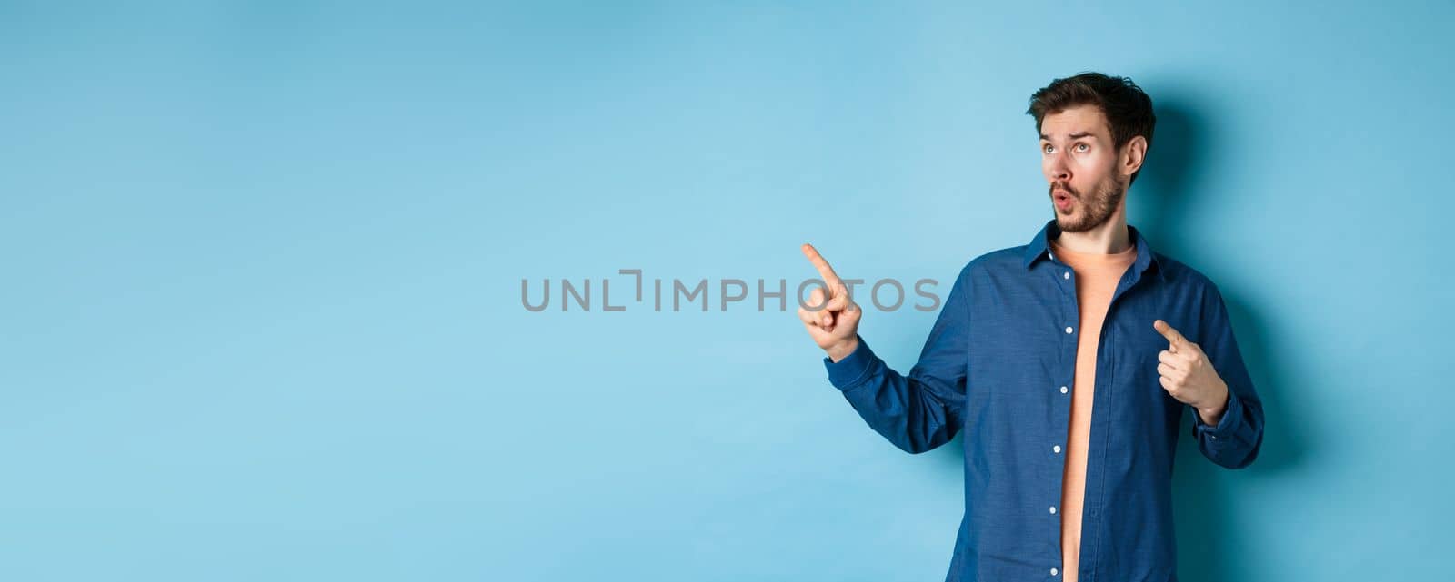 Impressed guy looking and pointing at upper left corner, showing advertisement, standing on blue background.