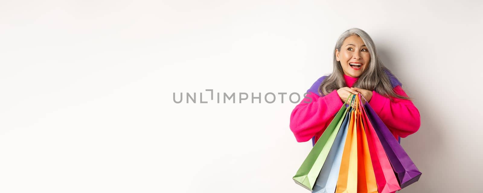 Fashionable senior asian woman shopaholic, hugging shopping bags and smiling joyful, buying with discounts, standing over white background.