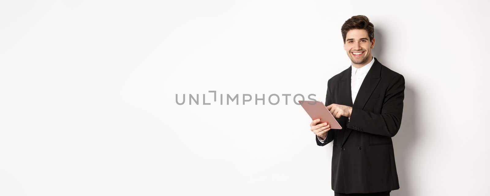 Image of handsome businessman in black suit, using digital tablet and smiling, standing against white background by Benzoix