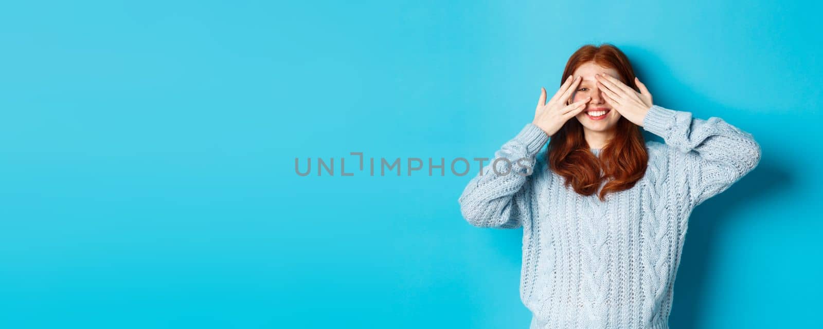 Excited teenage redhead girl open eyes to see holiday surprise, receiving presents, looking amazed at camera, standing over blue background by Benzoix