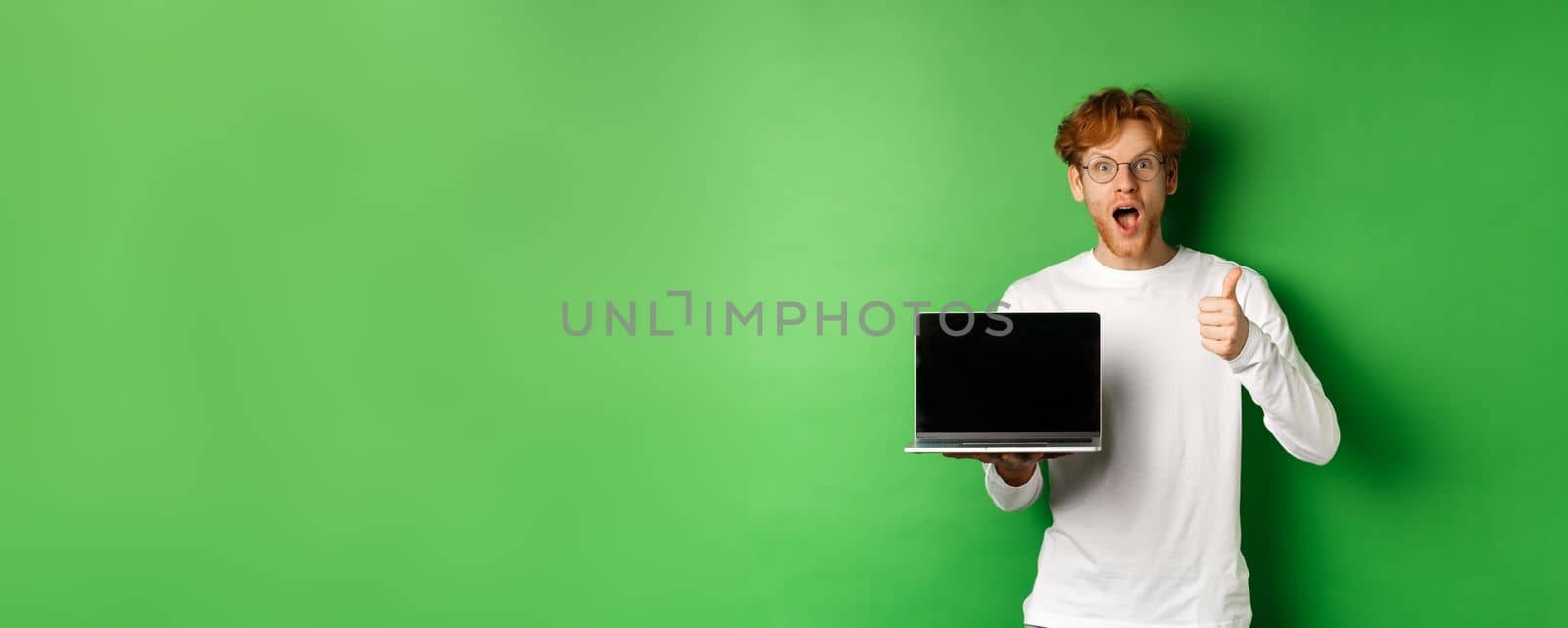 Impressed caucasian man with red hair and beard, showing blank laptop screen and thumb-up, looking amazed at camera, praising something online, green background by Benzoix