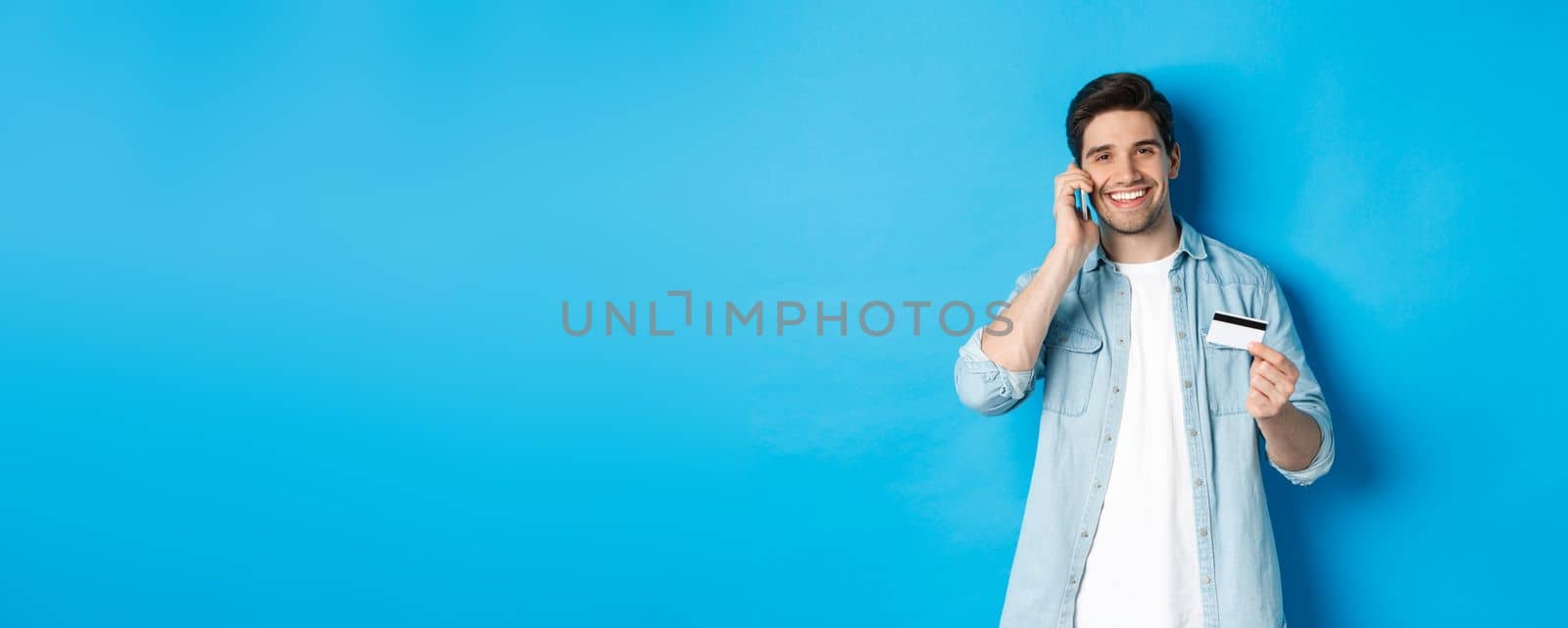 Handsome man calling bank and holding credit card, having mobile conversation, standing over blue background.