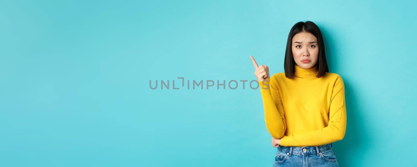 Shopping concept. Sad and gloomy asian girl in yellow sweater pointing finger left, frowning and showing bad news on copy space, standing over blue background.