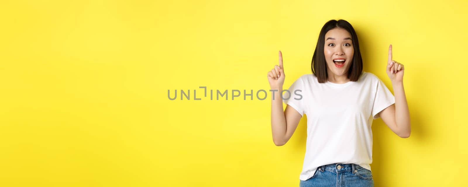 Beauty and fashion concept. Beautiful asian woman in white t-shirt pointing fingers up, standing over yellow background.