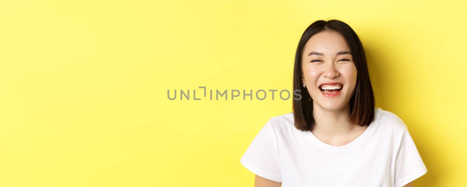 Close up of happy young woman having fun, smiling and laughing carefree, standing in white t-shirt over yellow background.