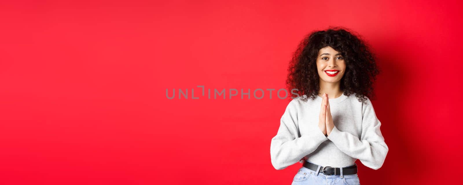Beautiful smiling italian woman saying thank you, holding hands in namaste gesture and looking at camera grateful, standing on red background.