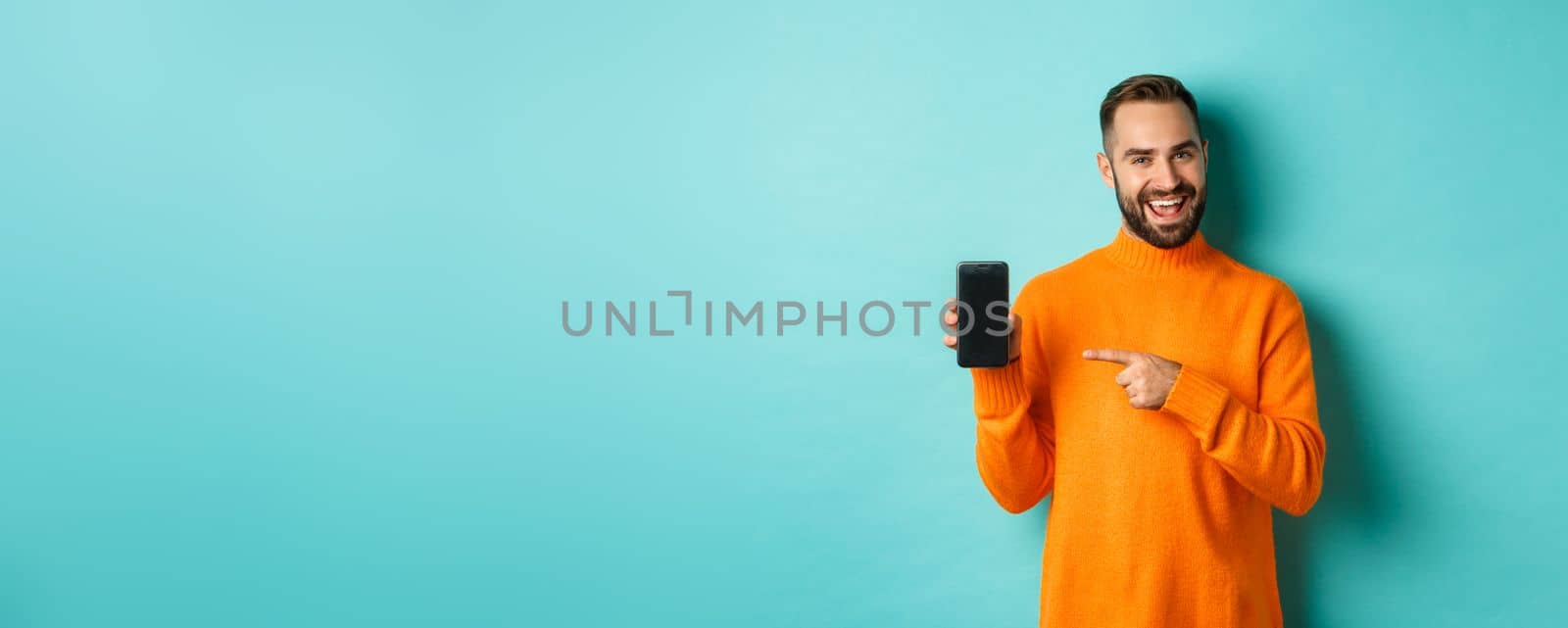 Handsome bearded man in orange sweater, pointing finger at mobile phone screen, showing application smartphone, standing over light blue background by Benzoix