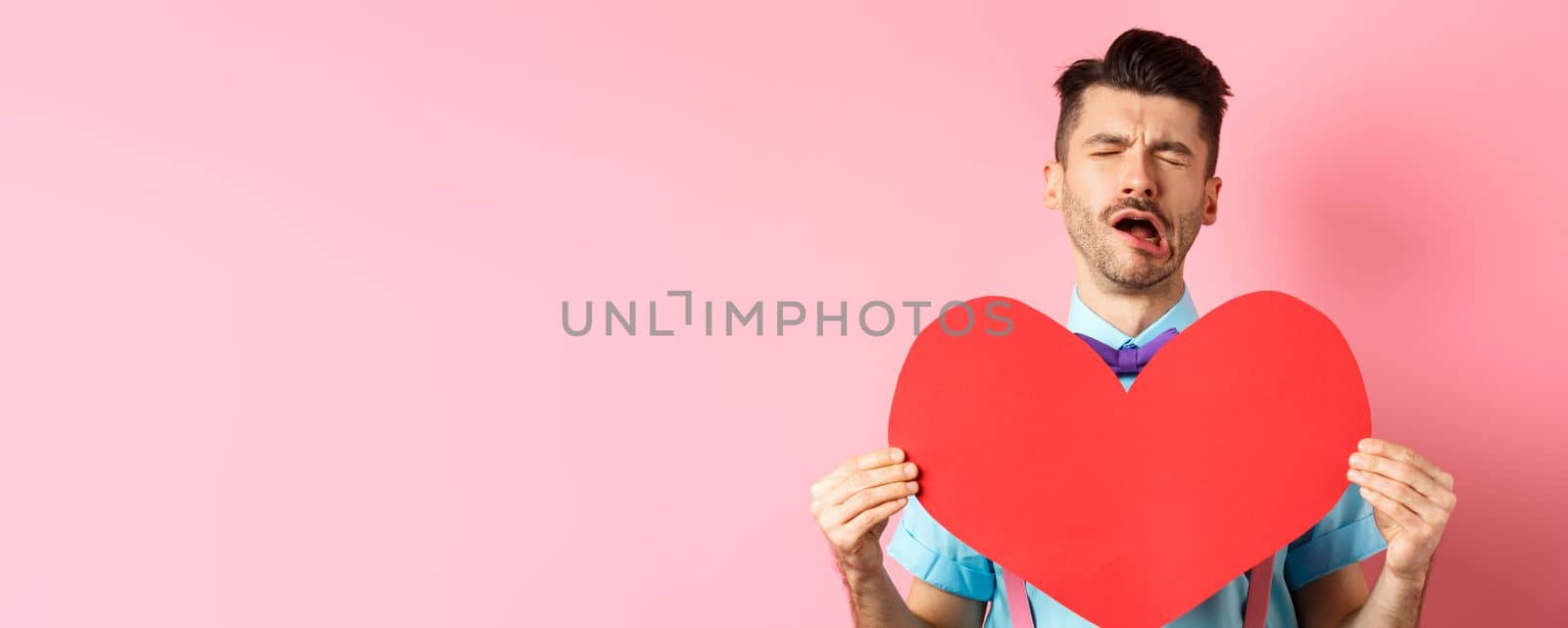 Valentines day concept. Sad and lonely man feeling heartbroken, being rejected, showing big red heart cutout and crying from break-up, standing on pink background.