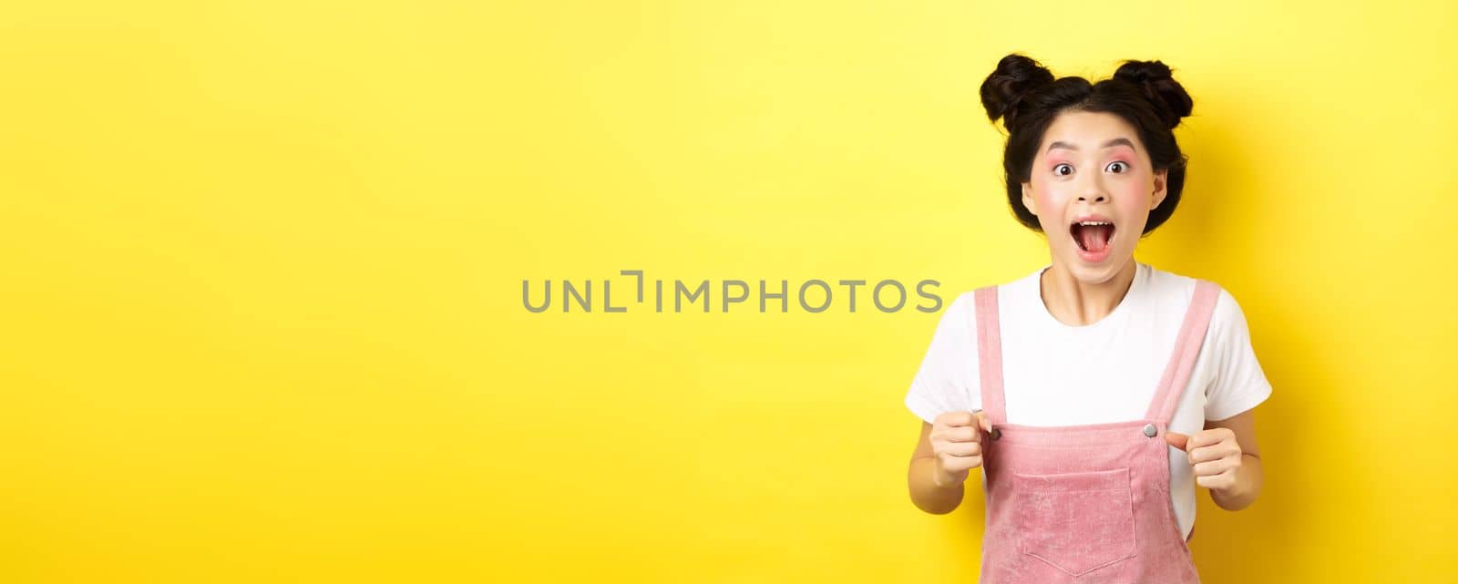 Excited asian woman open mouth, looking fascinated and amazed, receive good news, standing in summer clothes on yellow background.