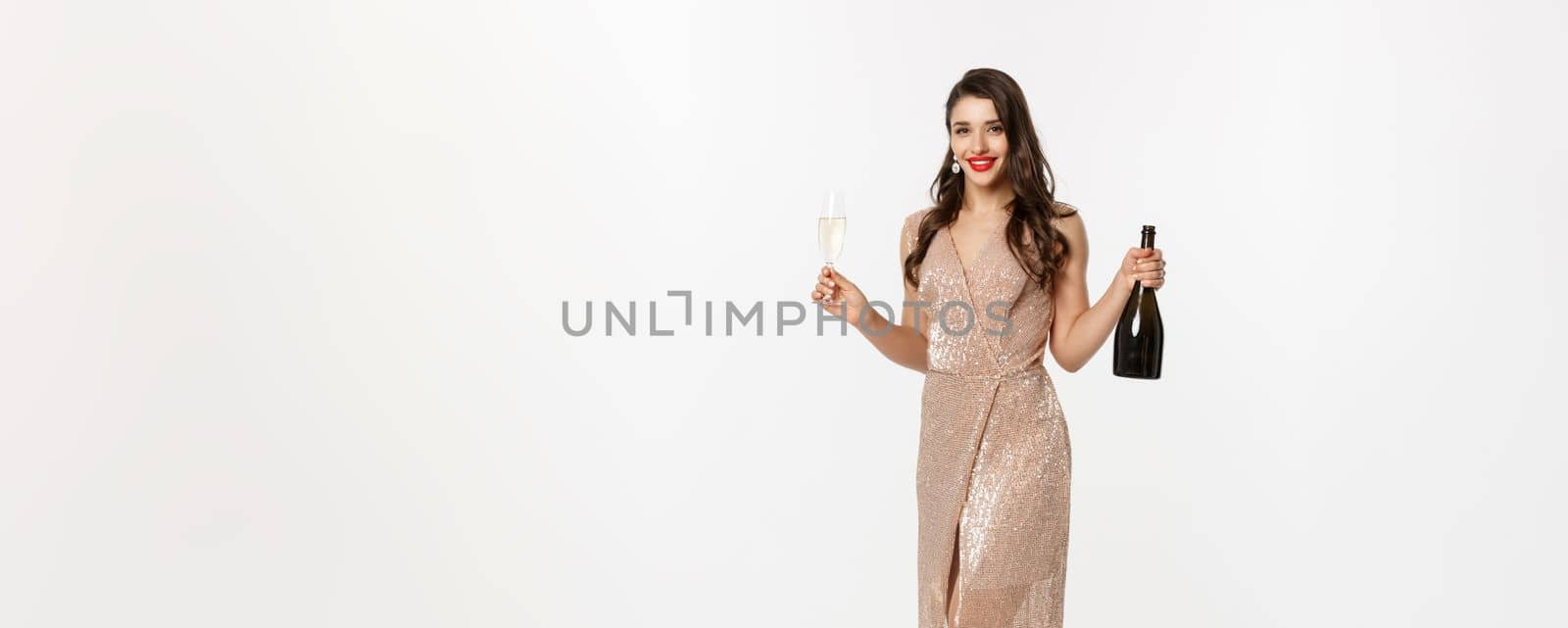 Christmas party and celebration concept. Full length of elegant woman with red lips, luxury dress, holding glass of champagne and bottle, white background.