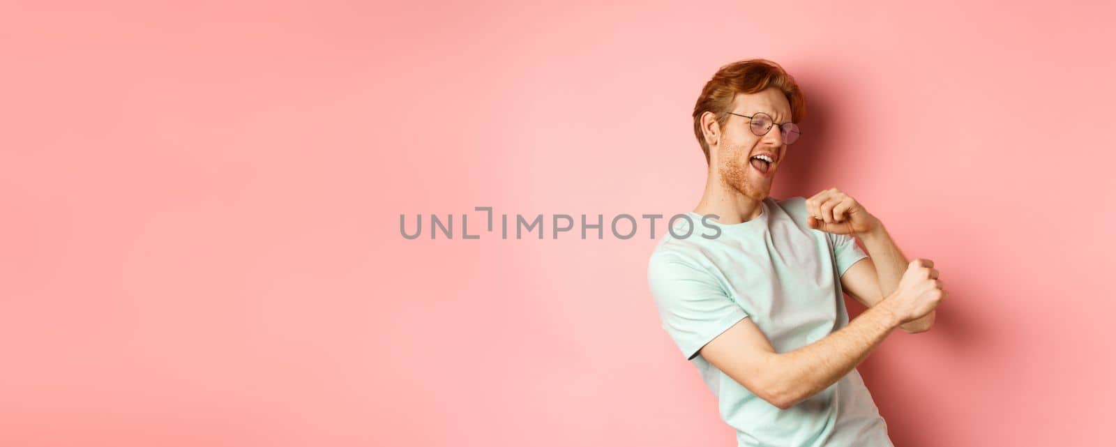 Summer vacation concept. Happy young man with red hair dancing and having fun, singing along music, standing over pink background.