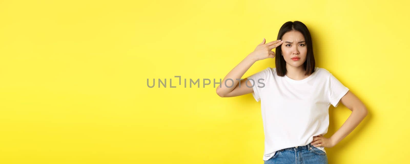 Woman showing kill me please gesture, shooting herself in head with finger gun from boredom, standing upset over yellow background.