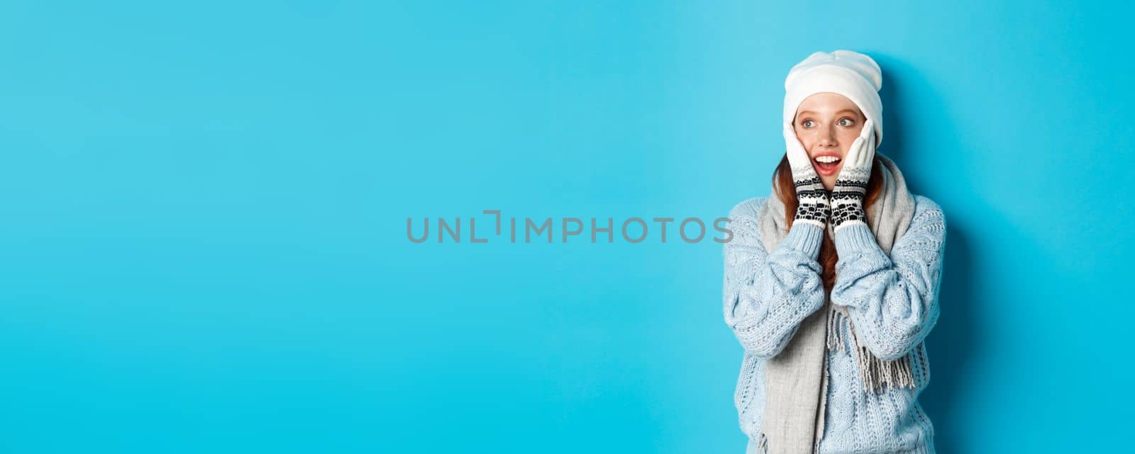 Excited redhead girl staring left at logo, wearing winter clothes, beanie, gloves and sweater, standing over blue background by Benzoix