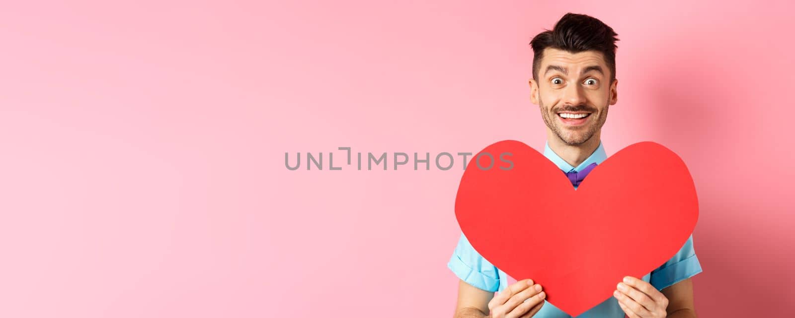 Hopeful man in love showing red heart sign, smiling at camera, waiting for soulmate on Valentines day, standing on pink background by Benzoix