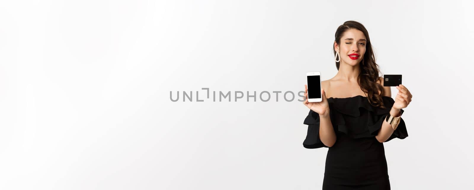 Fashion and shopping concept. Beautiful woman with red lips, winking at camera, showing smartphone screen and credit card, buying online, white background by Benzoix