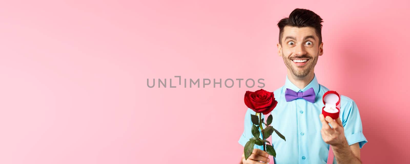 Valentines day. Smiling handsome man asking to marry him, showing engagement ring and red rose, standing romantic on pink background by Benzoix