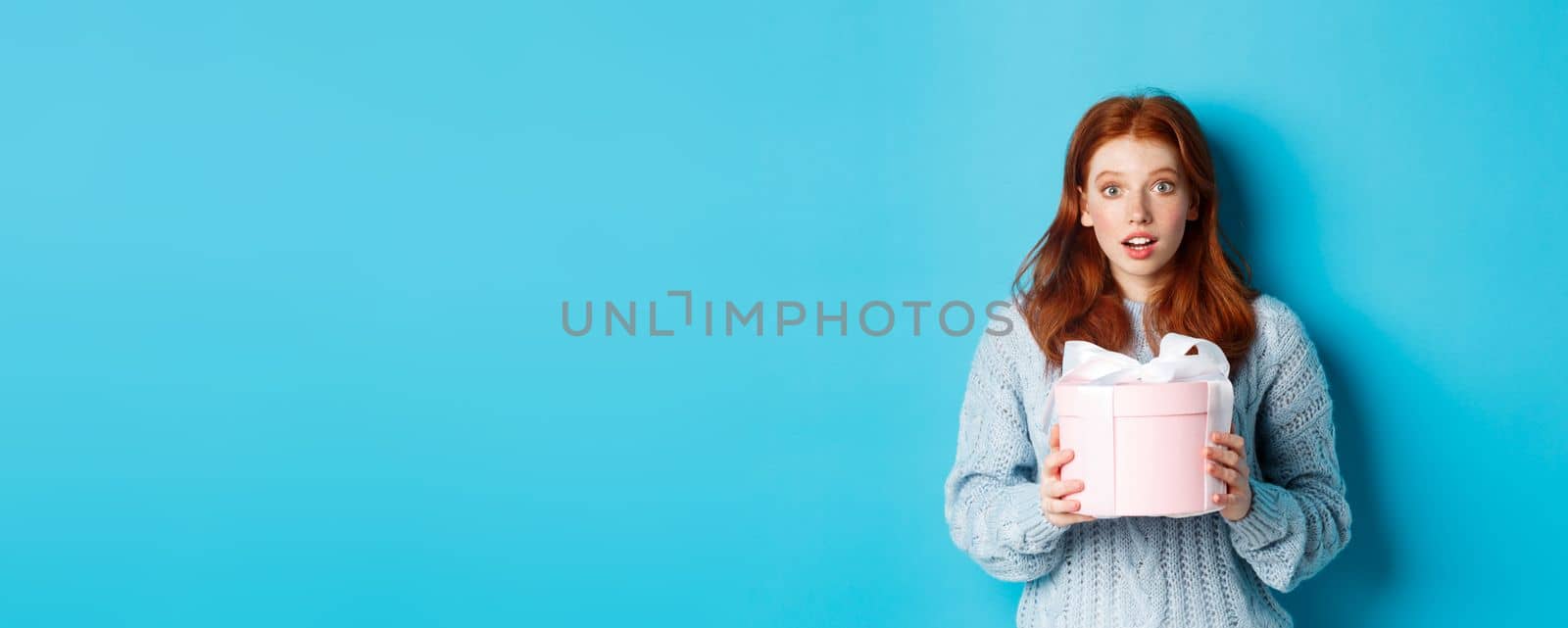 Surprised redhead girl receiving valentines gift, holding box with present and staring at camera amazed, wearing sweater, standing over blue background by Benzoix