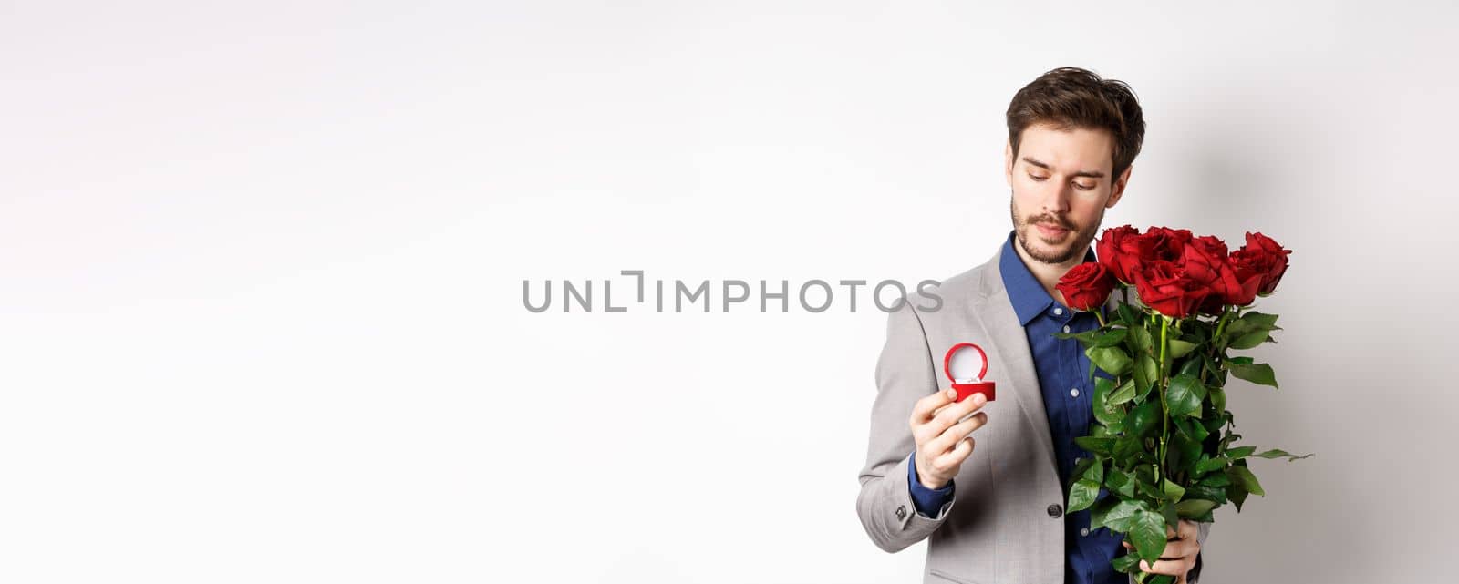 Romantic man in suit looking pensive at engagement ring, going to make a marriage proposal on Valentines day, holding bouquet of roses, standing over white background by Benzoix