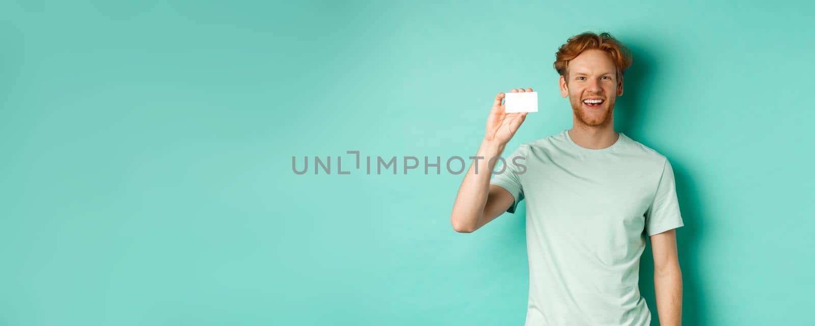 Shopping concept. Cheerful young man in t-shirt showing plastic credit card and smiling, standing over mint background.