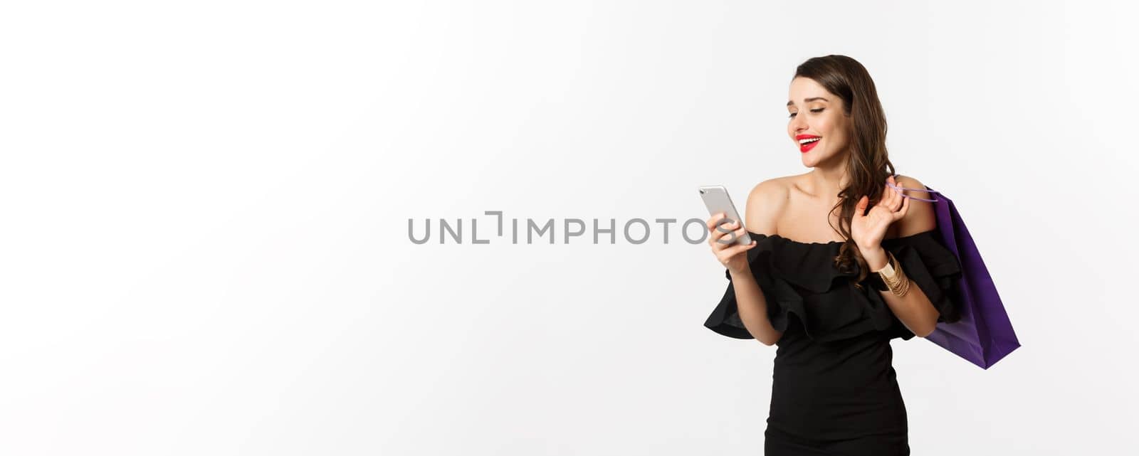 Beauty and shopping concept. Gorgeous woman in elegant black dress and makeup, order online on smartphone, holding bag and smiling, standing over white background by Benzoix