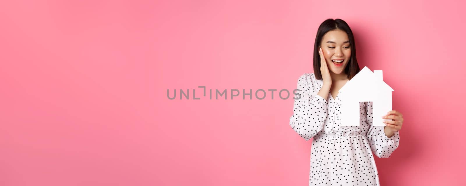 Real estate. Cute asian woman searching for apartment, looking happy at paper house model, standing against pink background.
