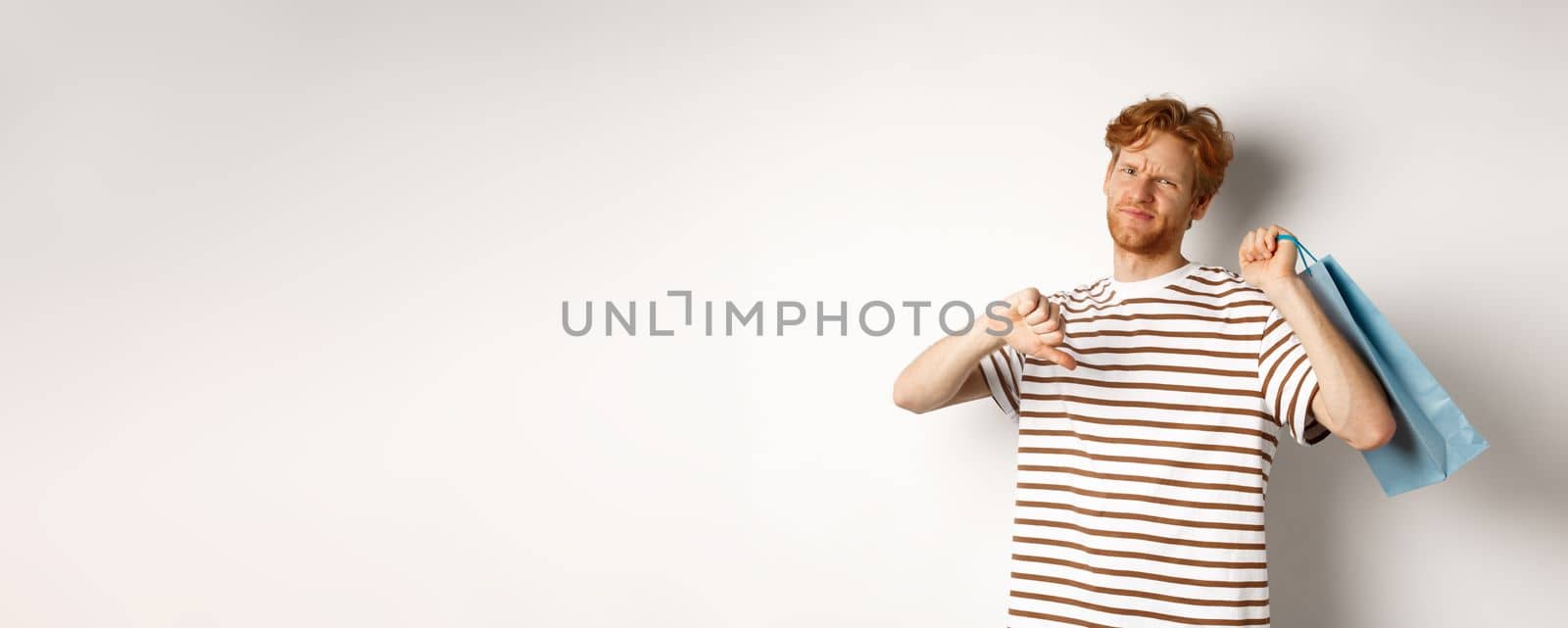 Disappointed young man with red hair and beard showing thumbs-down after bad shopping experience, holding bag over shoulder and frowning upset, white background.