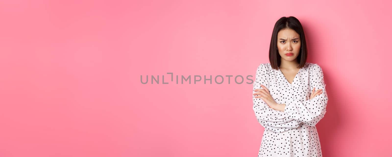 Timid and offended asian girl cross arms on chest, staring defensive and insulted at camera, standing in dress over pink background.