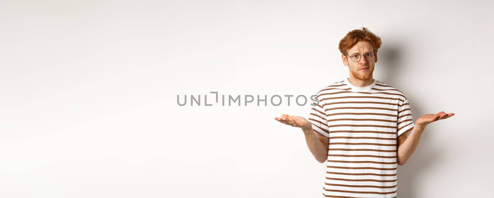 Confused boyfriend with red hair shrugging, spread hands sideways and staring at camera puzzled, asking what, stnading over white background.