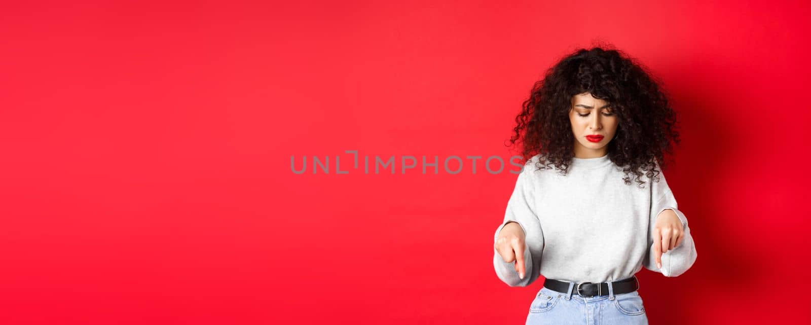 Worried young woman with curly hairstyle, looking down and pointing at empty space with concerned hesitant face, standing on red background.