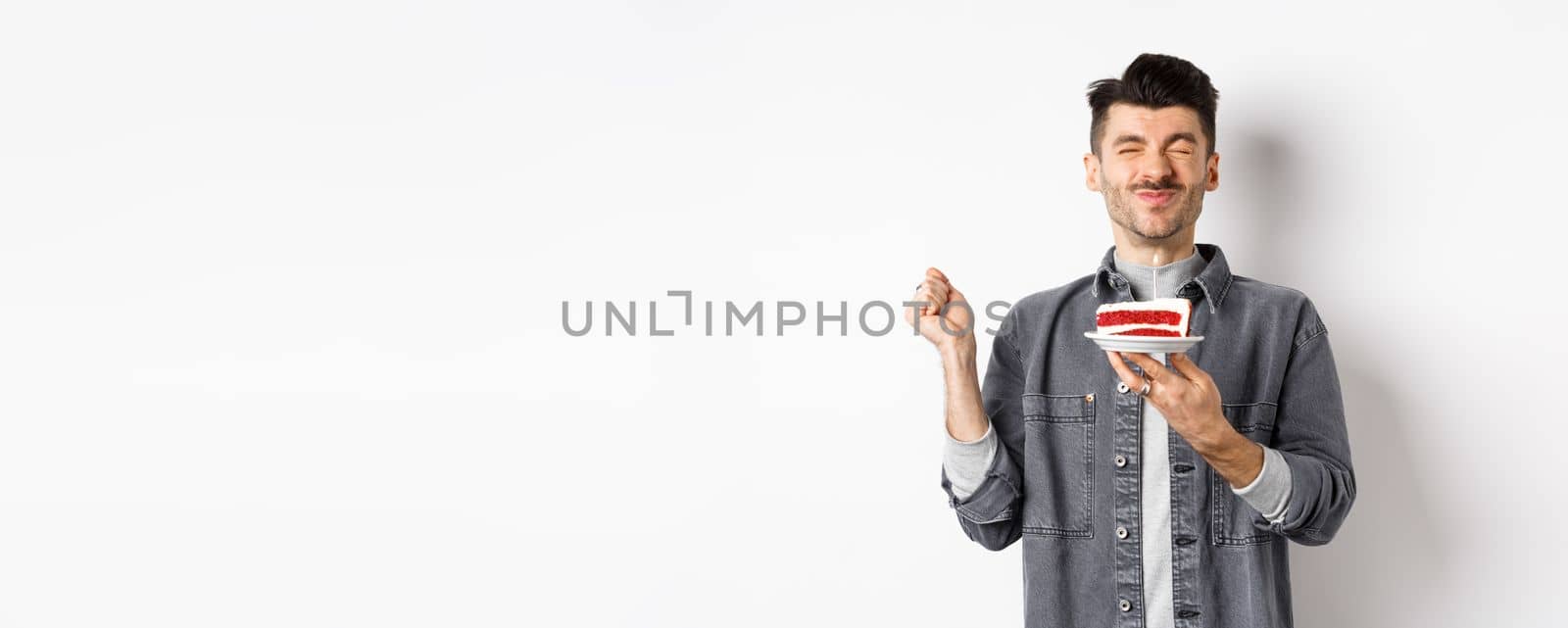 Happy birthday guy making wish on cake with candle, celebrating bday, standing on white background by Benzoix