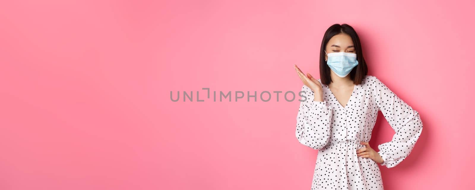 Covid-19, quarantine and lifestyle concept. Beautiful and confident asian woman in dress and face mask looking self-assured, standing sassy against pink background.