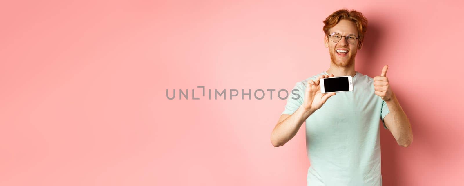 Happy european man with ginger hair and glasses, showing mobile blank screen horizontally and thumbs-up, recommend online promo, pink background.
