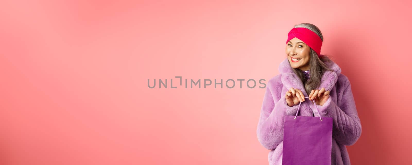 Shopping and fashion concept. Fashionable gradmother bought present and smiling, holding purple bag in hands, standing over pink background by Benzoix