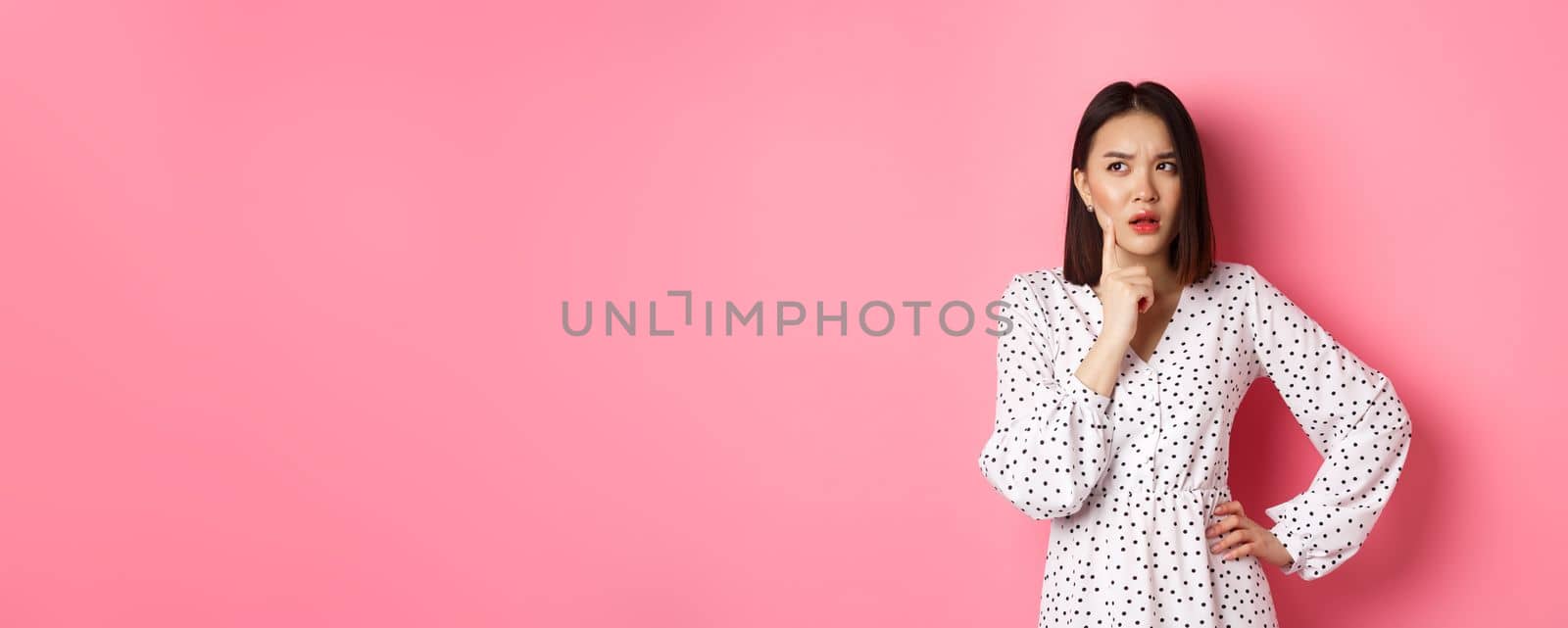 Thoughtful asian woman making assumption, looking up and thinking, deciding something, standing in dress over pink background.