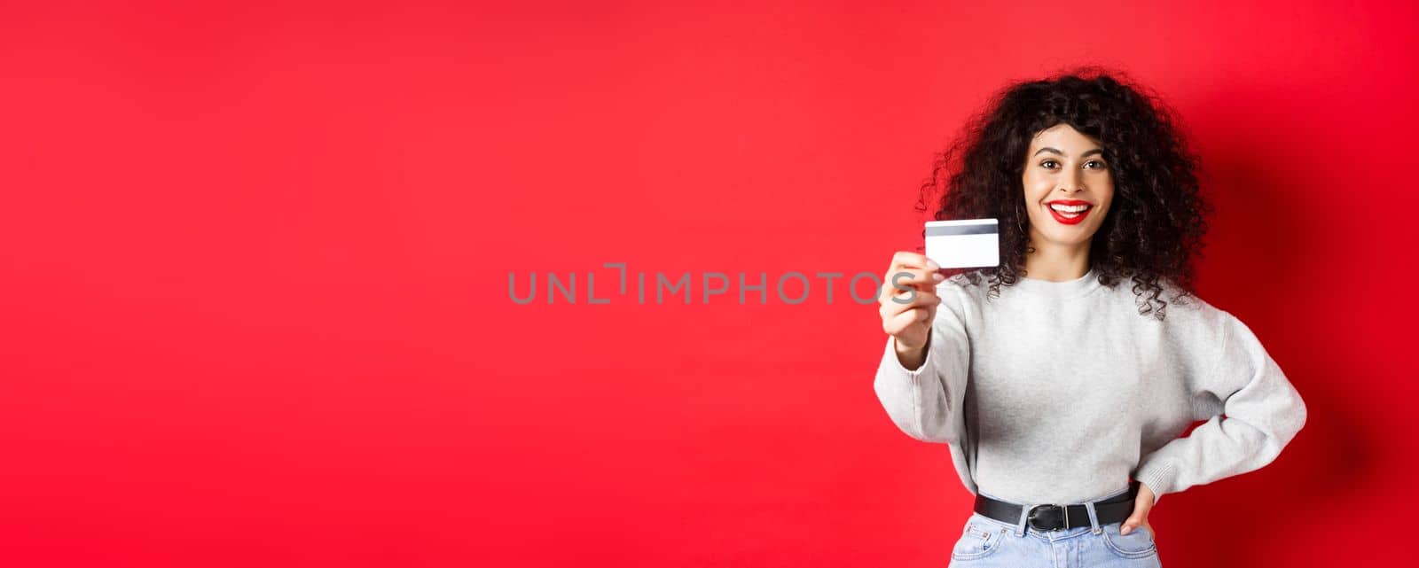 Image of modern woman with curly hair, extending hand and showing plastic credit card, recommending bank or shopping offer, red background by Benzoix